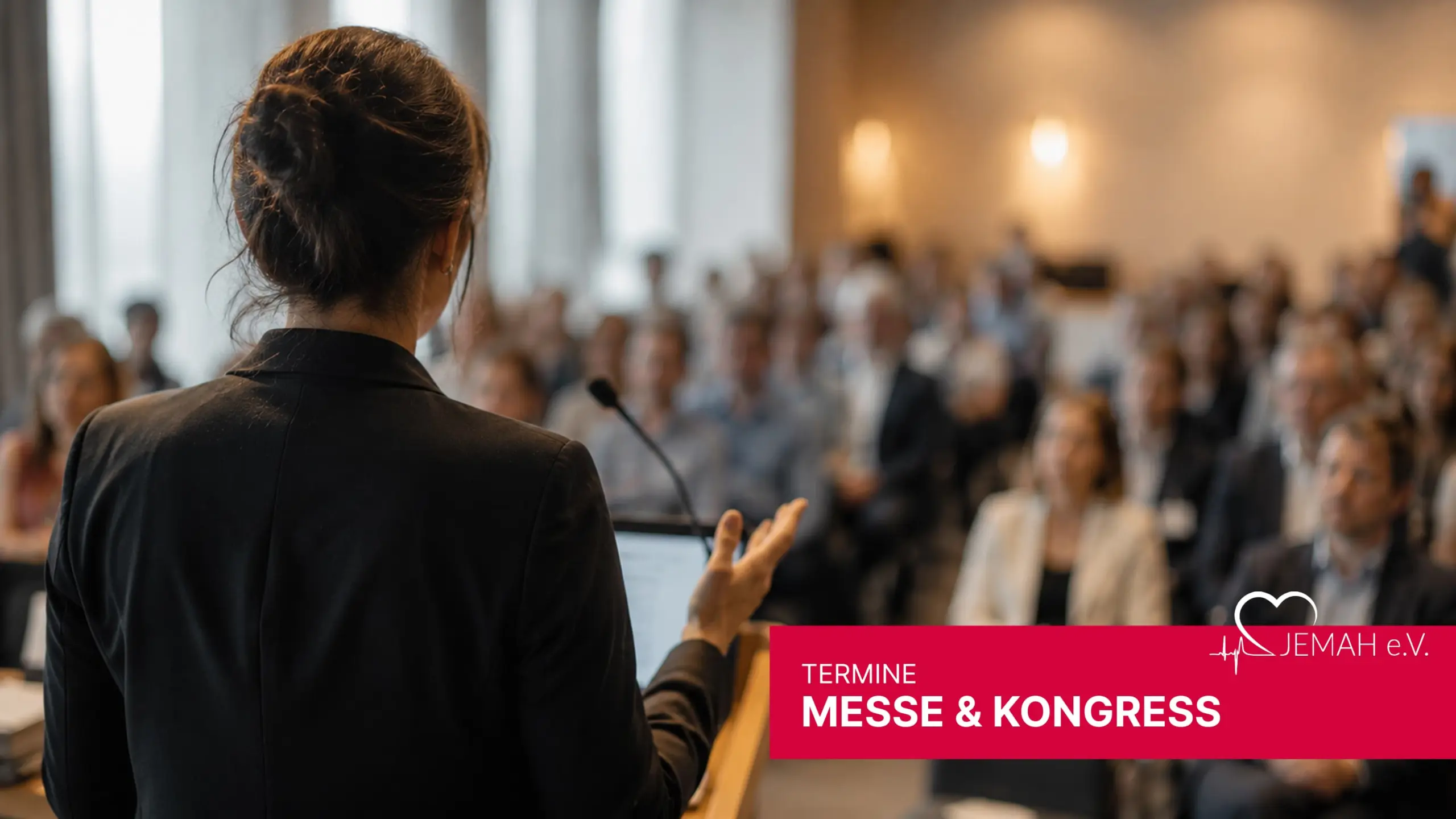 Referentin bei einer jemah Messe & Kongress Veranstaltung spricht vor Publikum an einem Pult mit Mikrofon.