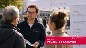 Ein jemah e.V. Mitarbeiter in blauer Fleecejacke berät zwei Besucher an einem Infostand im Freien, im Hintergrund ein jemah Banner.