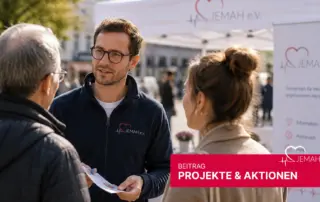Ein jemah e.V. Mitarbeiter in blauer Fleecejacke berät zwei Besucher an einem Infostand im Freien, im Hintergrund ein jemah Banner.